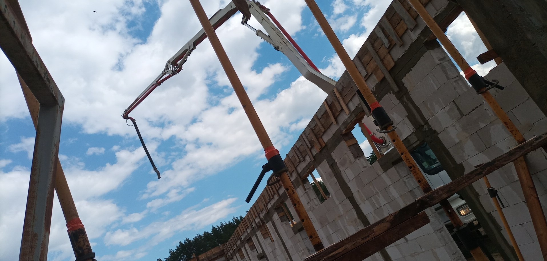 Budowa domu z betonu komórkowego widoczna z dołu, z użyciem pompy do betonu i podpór szalunkowych, na tle błękitnego, pochmurnego nieba.