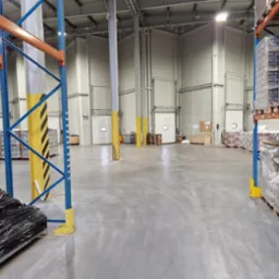 Wnętrze magazynu z regałami wypełnionymi zapakowanymi towarami na paletach, widoczne kolumny z żółto-czarnymi pasami ostrzegawczymi.