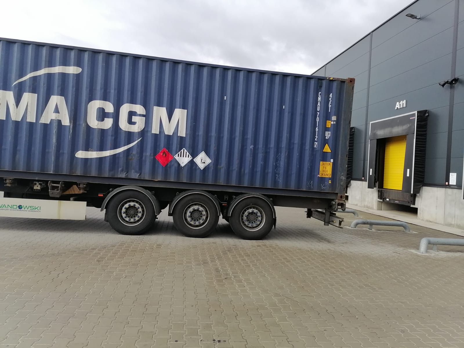 Niebieski kontener CMA CGM na naczepie ciężarówki, z widocznymi oznaczeniami ostrzegawczymi, zaparkowany przy rampie magazynowej z żółtymi drzwiami.