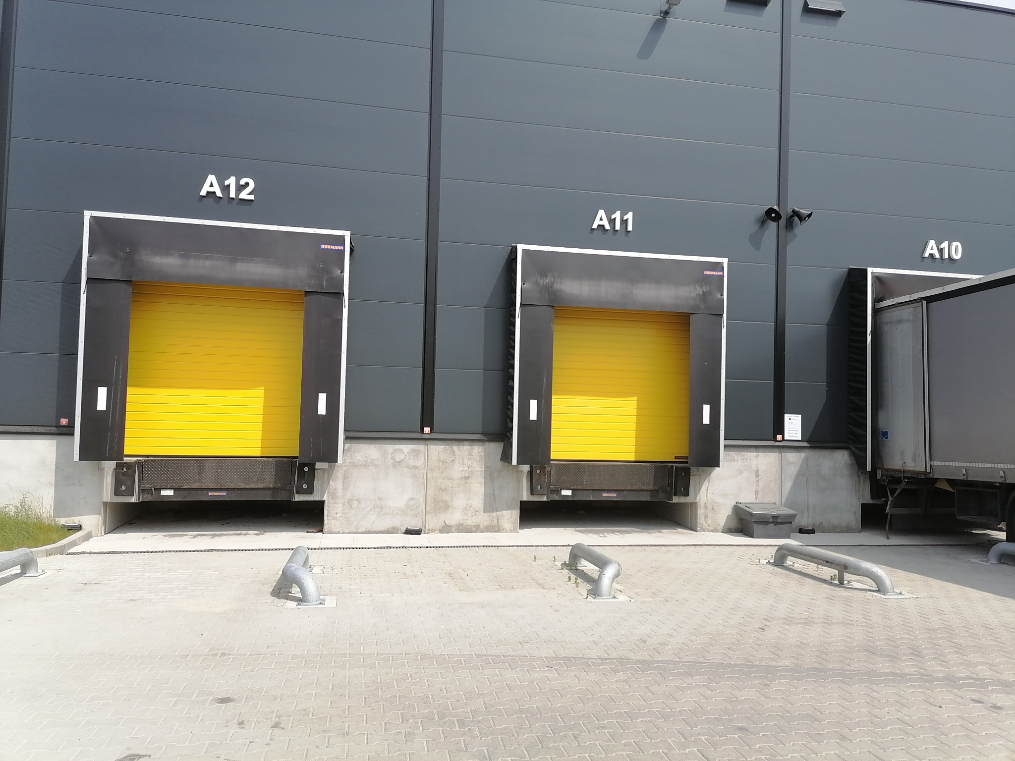 Rampa magazynowa z trzema żółtymi bramami segmentowymi, oznaczonymi A12, A11 i A10, z częściowo widoczną naczepą ciężarówki przy bramie A10.