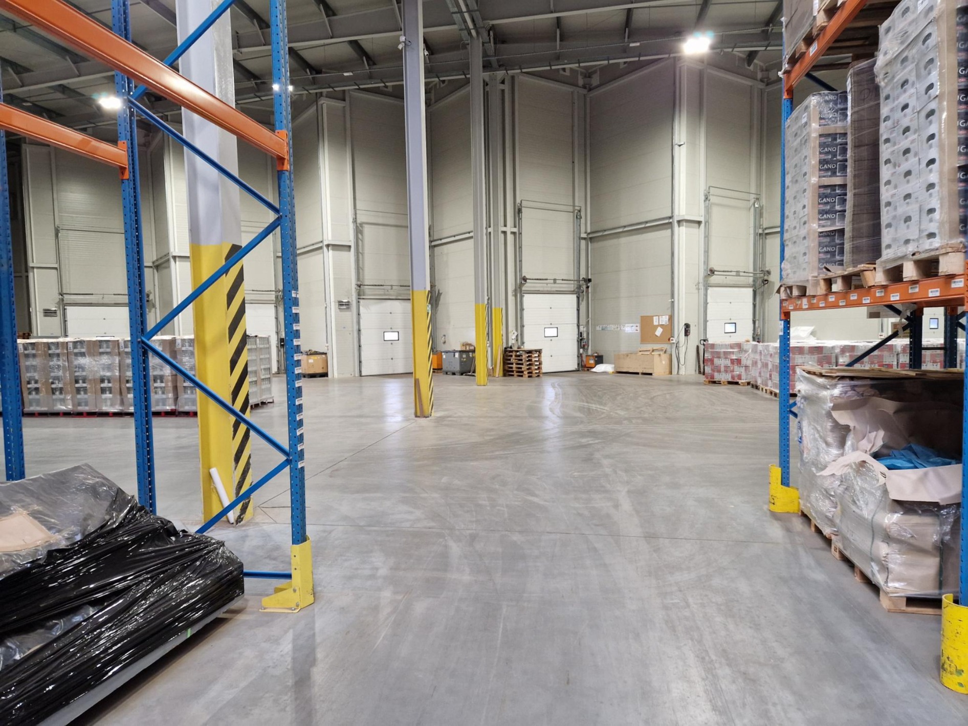 Wnętrze magazynu z regałami wypełnionymi zapakowanymi towarami na paletach, widoczne kolumny z żółto-czarnymi pasami ostrzegawczymi.