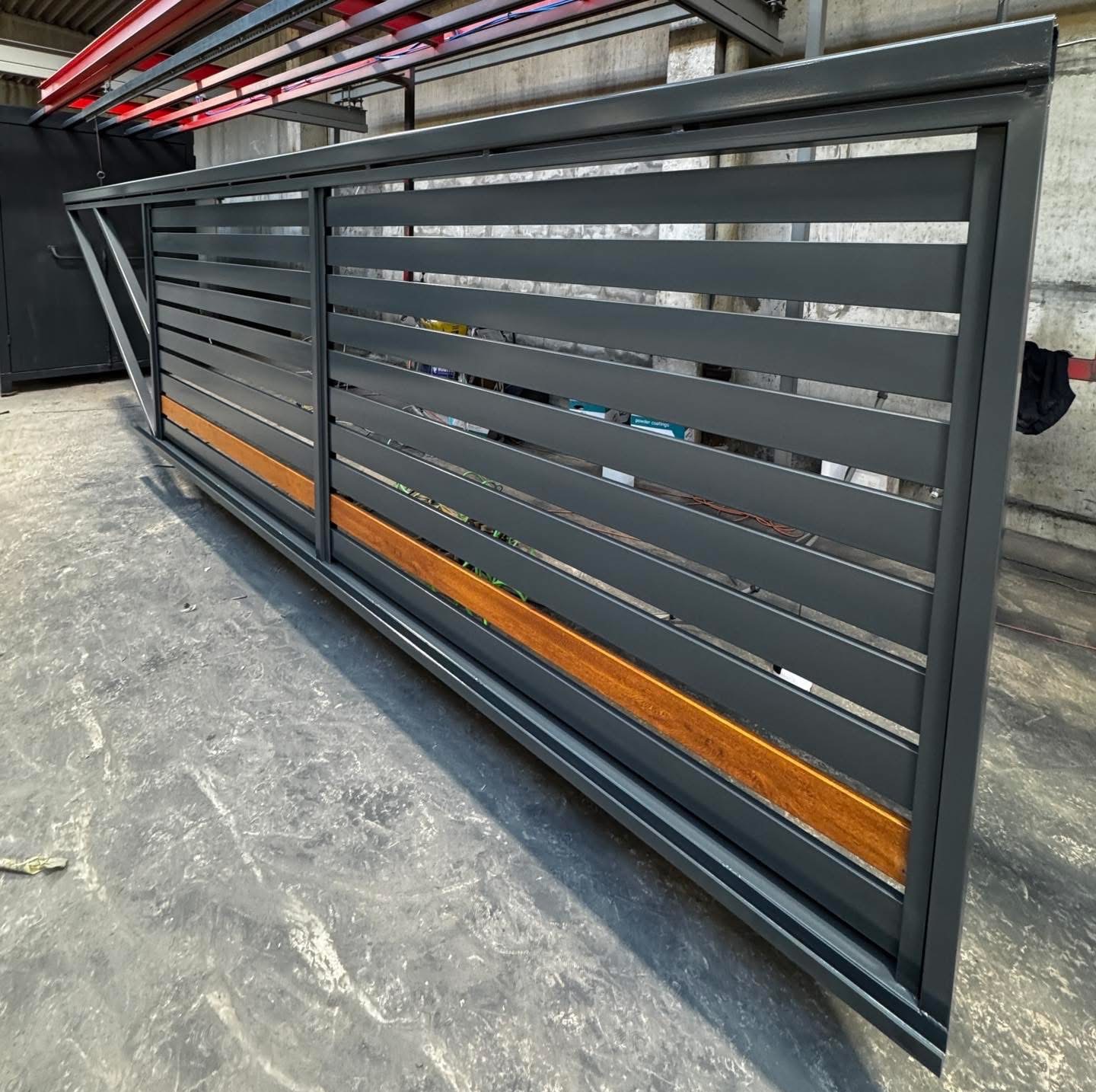 Nowoczesna, przesuwna brama metalowa w odcieniach szarości z drewnianymi akcentami, widoczna w hali produkcyjnej. Minimalistyczny design, poziome lamele.