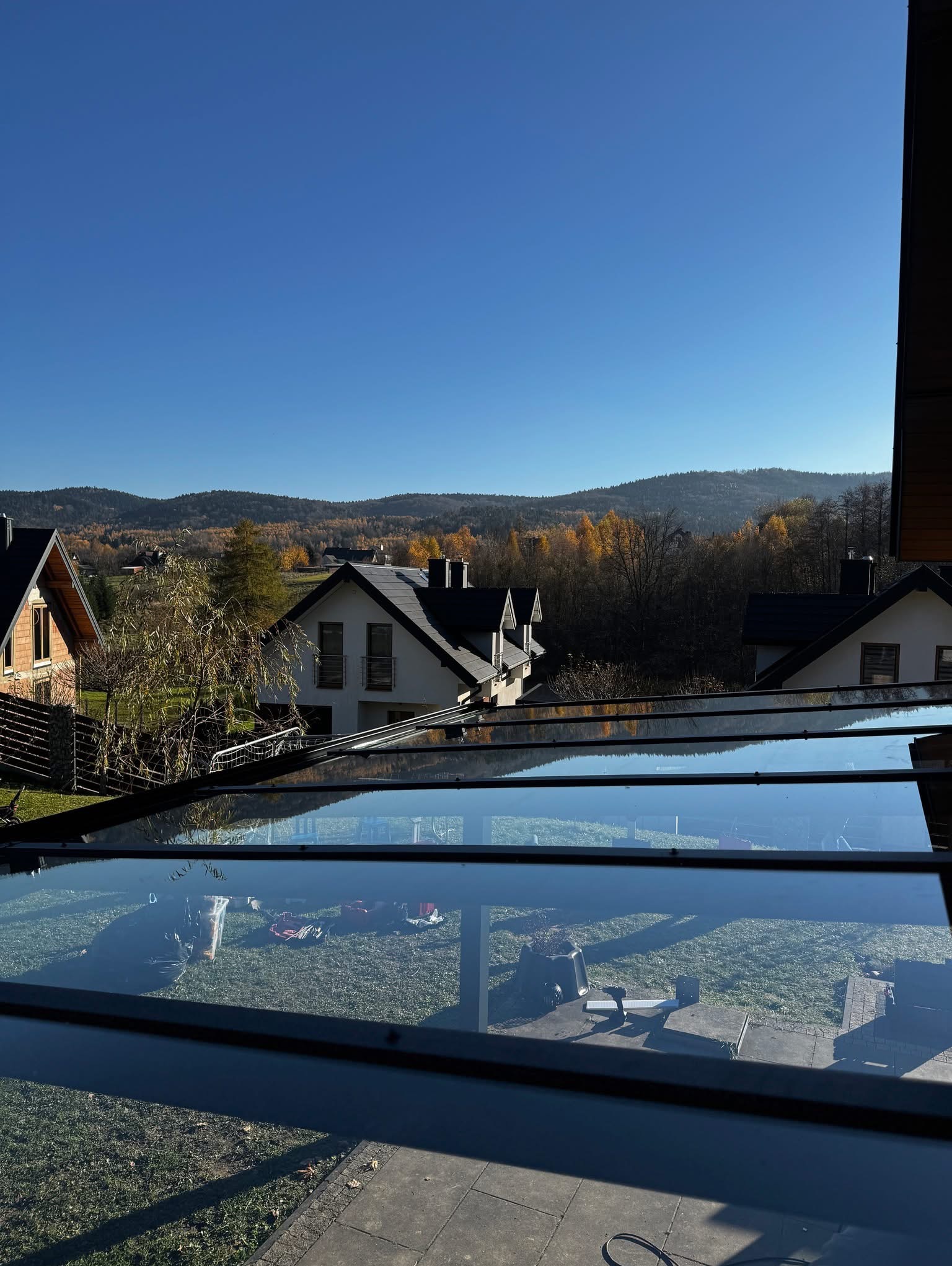 Przeszklona zabudowa balkonu z widokiem na górzysty krajobraz w jesiennej scenerii. Widoczne narzędzia i akcesoria montażowe na trawie.