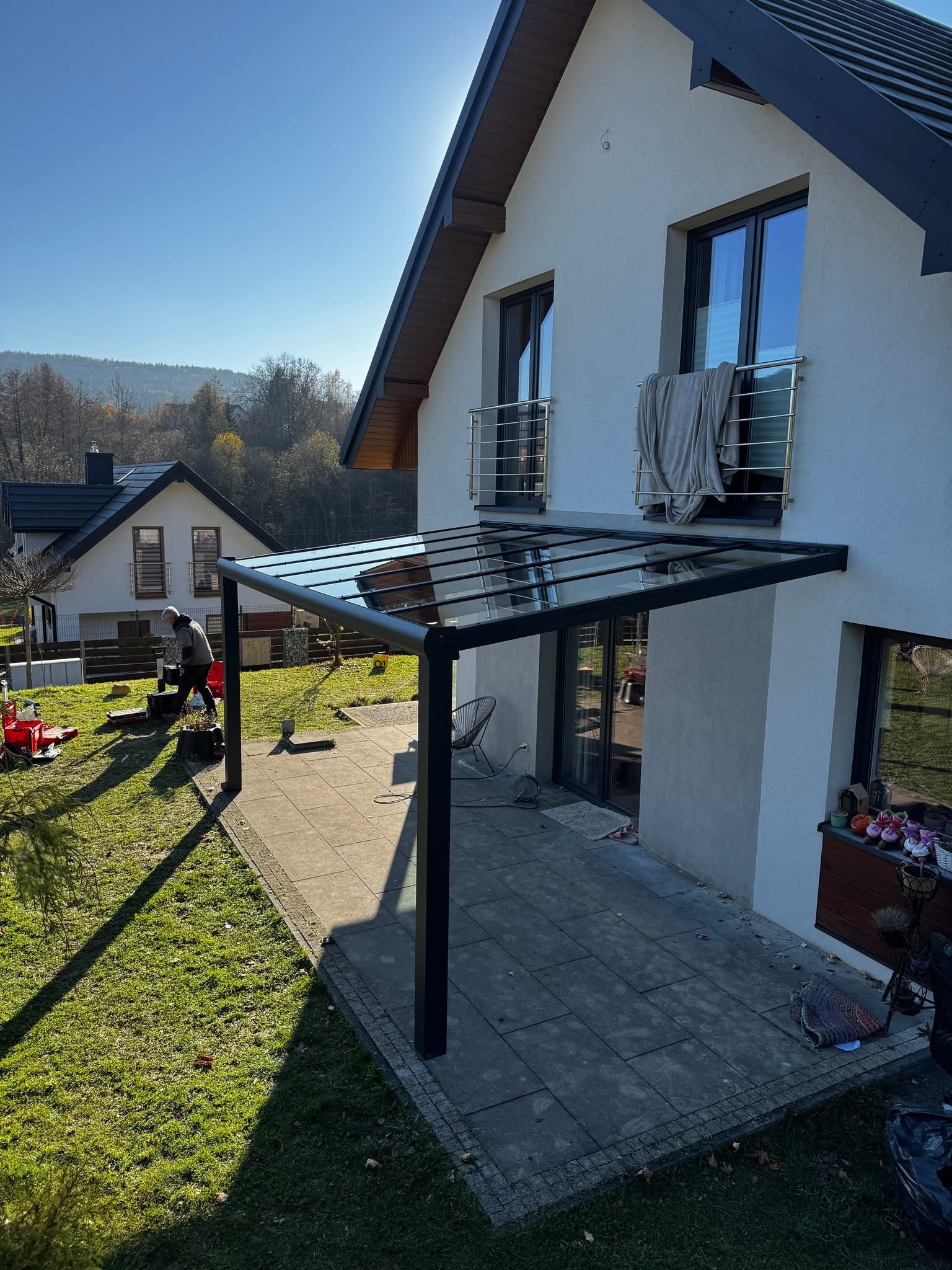 Nowoczesna pergola metalowa z przezroczystym zadaszeniem, przylegająca do jasnego domu. Widoczny fragment ogrodu i pracownik w oddali. Słoneczny dzień.
