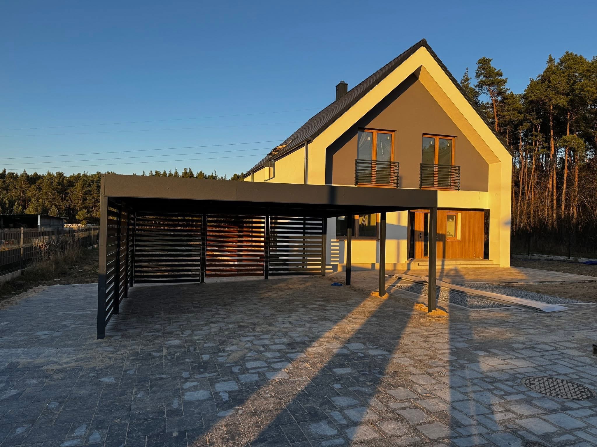 Nowoczesna, grafitowa pergola metalowa przy domu jednorodzinnym z szarej kostki brukowej, oświetlona ciepłym światłem zachodzącego słońca.