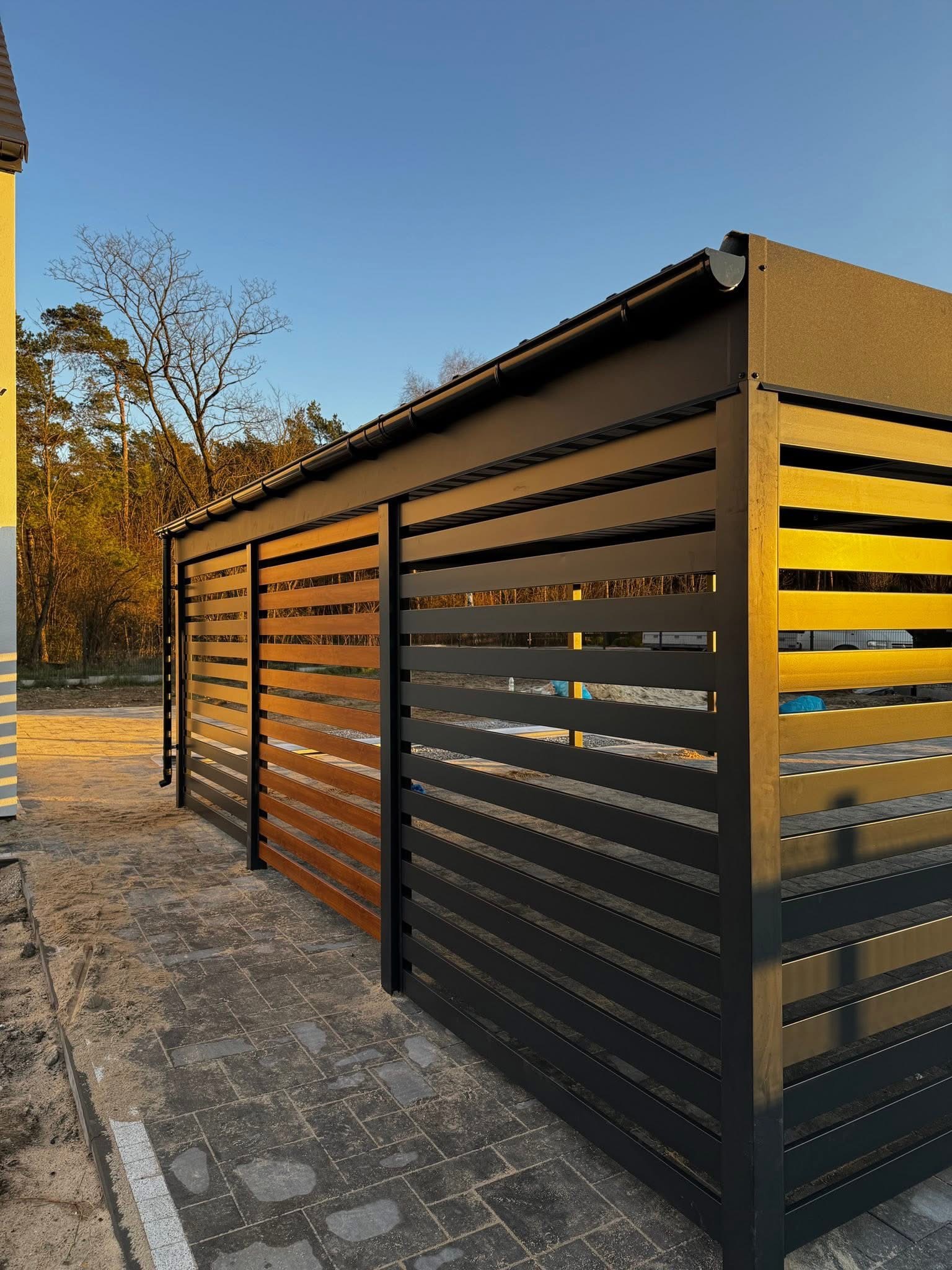 Nowoczesna pergola metalowa z poziomymi lamelami w kolorze grafitowym i drewnopodobnym, zadaszona, z rynną, na tle błękitnego nieba i drzew.