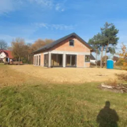 Surowy stan nowo budowanego domu z czerwonej cegły z dużymi oknami, widoczne fundamenty i teren przygotowany pod trawnik, niebieski przenośny WC w tle.