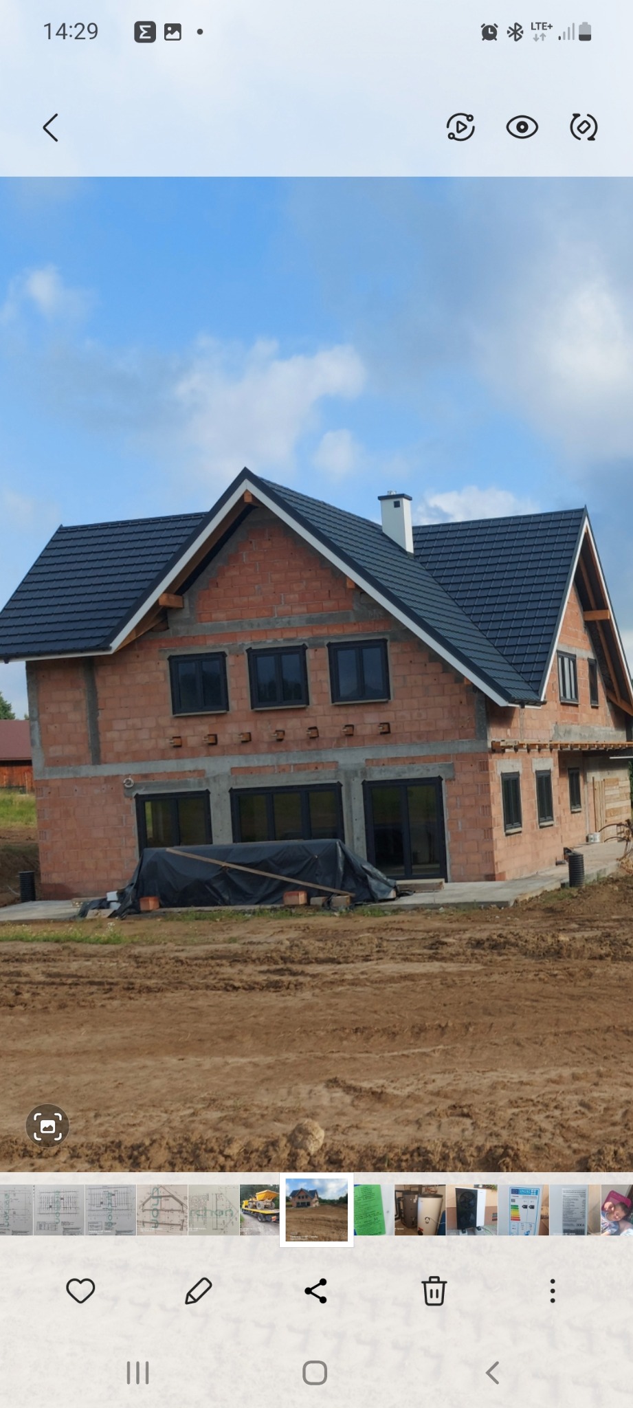 Surowy stan domu jednorodzinnego z czerwonej cegły z ciemnymi oknami i grafitowym dachem, widoczny na tle nieba i przygotowanego terenu.