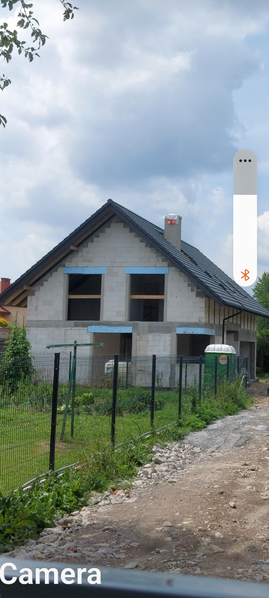 Dwukondygnacyjny dom murowany w stanie surowym zamkniętym z ciemną dachówką i oknami zabezpieczonymi niebieską pianką, widoczny zza metalowego ogrodzenia i przenośnej toalety.