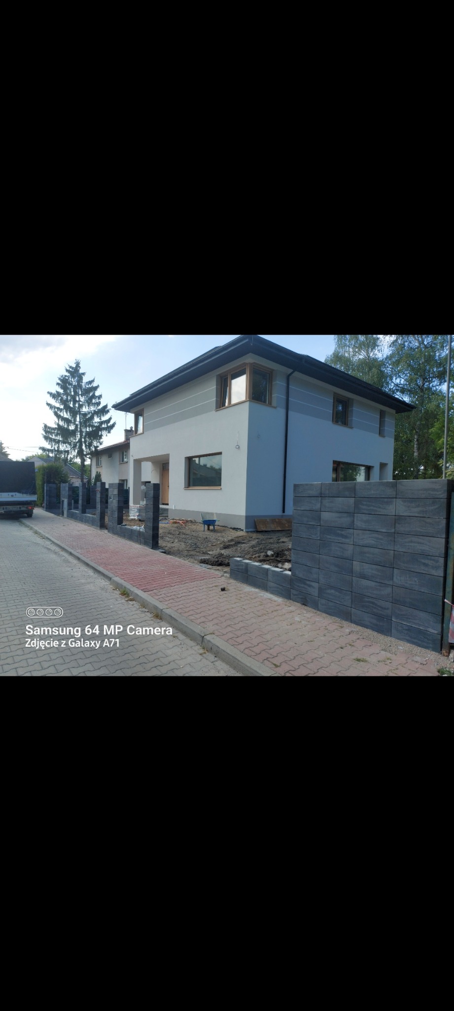 Dwukondygnacyjny dom w jasnych kolorach z szarymi akcentami, widoczny z zewnątrz, z nowo postawionym ogrodzeniem z szarych bloczków betonowych, kostka brukowa na chodniku i widoczne przygotowania...