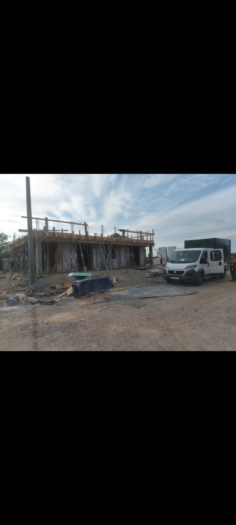 Budowa domu w trakcie realizacji z widocznymi ścianami, deskowaniem i podpartymi słupami, na placu budowy stoi biały samochód dostawczy.