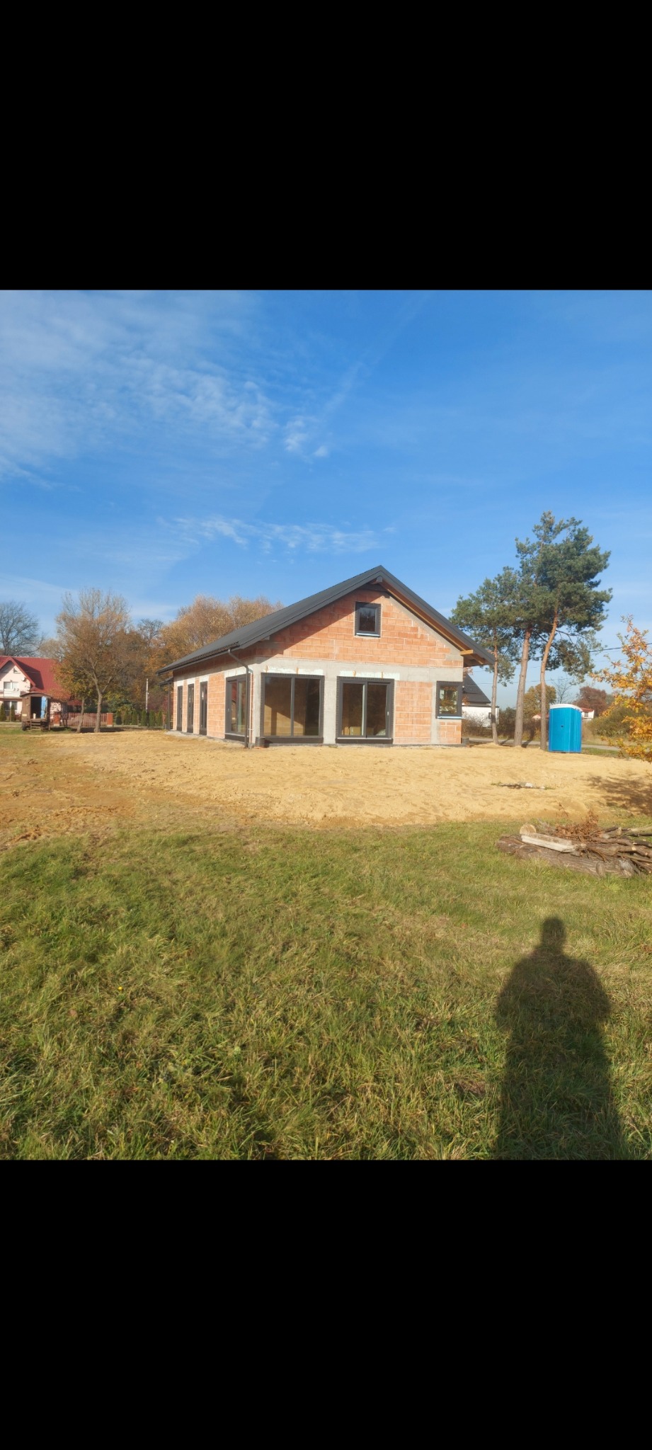 Surowy stan nowo budowanego domu z czerwonej cegły z dużymi oknami, widoczne fundamenty i teren przygotowany pod trawnik, niebieski przenośny WC w tle.