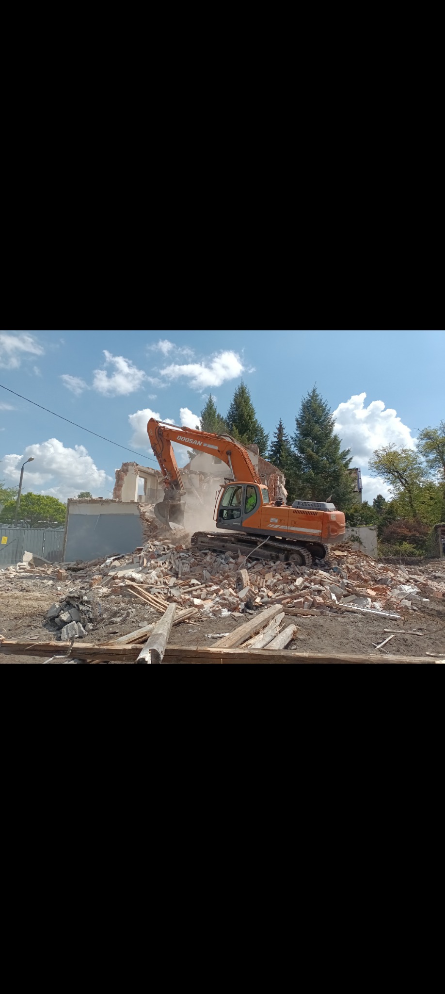 Pomarańczowa koparka Doosan na gruzowisku podczas rozbiórki budynku. W tle drzewa iglaste i błękitne niebo z chmurami.