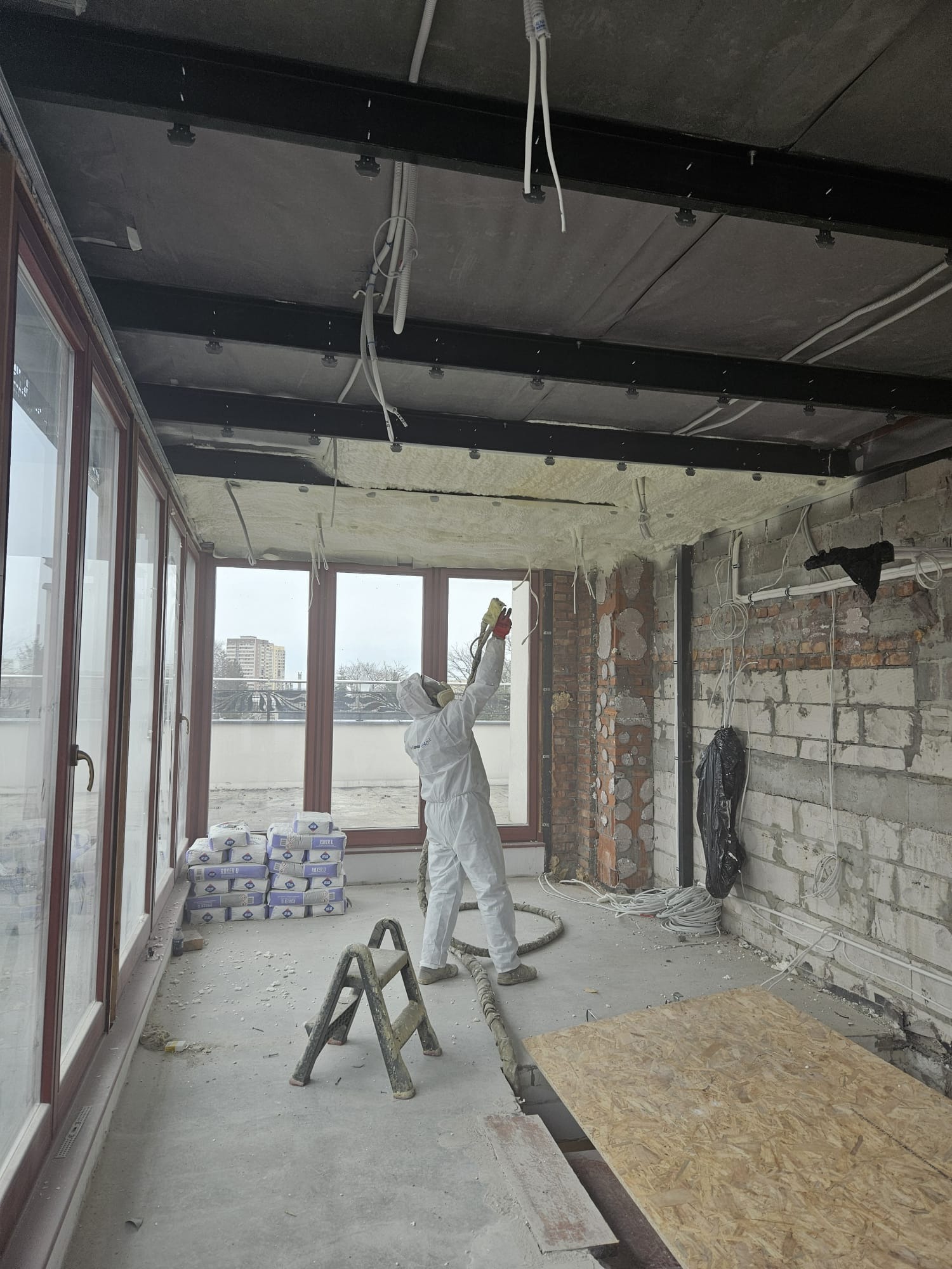 Pracownik w kombinezonie ochronnym natryskuje piankę PUR na sufit poddasza w trakcie ocieplania, widoczne elementy konstrukcji stalowej, okna balkonowe i worki z materiałem budowlanym.