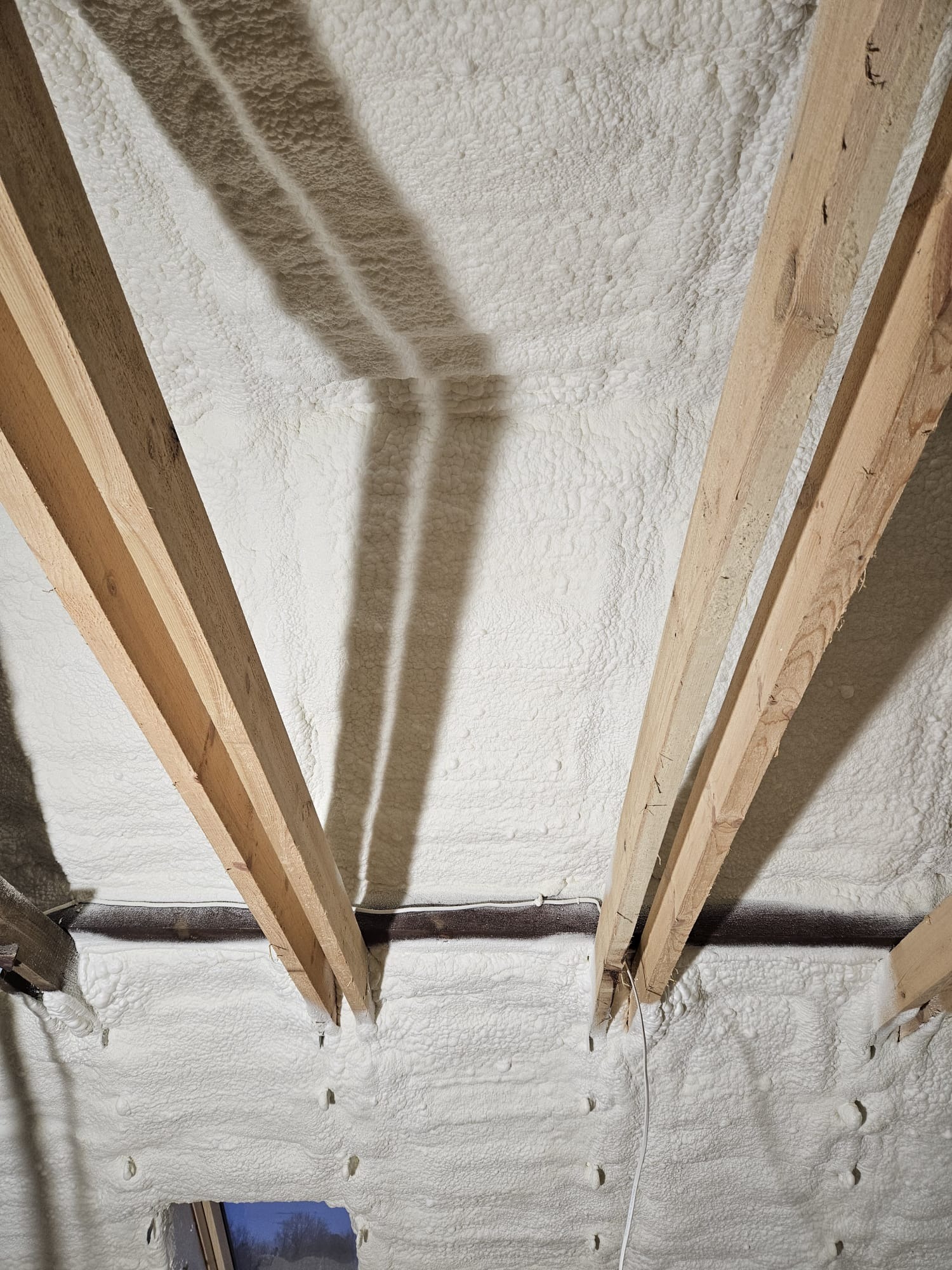 Poddasze z drewnianą konstrukcją krokwiową, pokryte grubą warstwą białej pianki izolacyjnej, widoczne cienie krokwi i fragment okna dachowego.