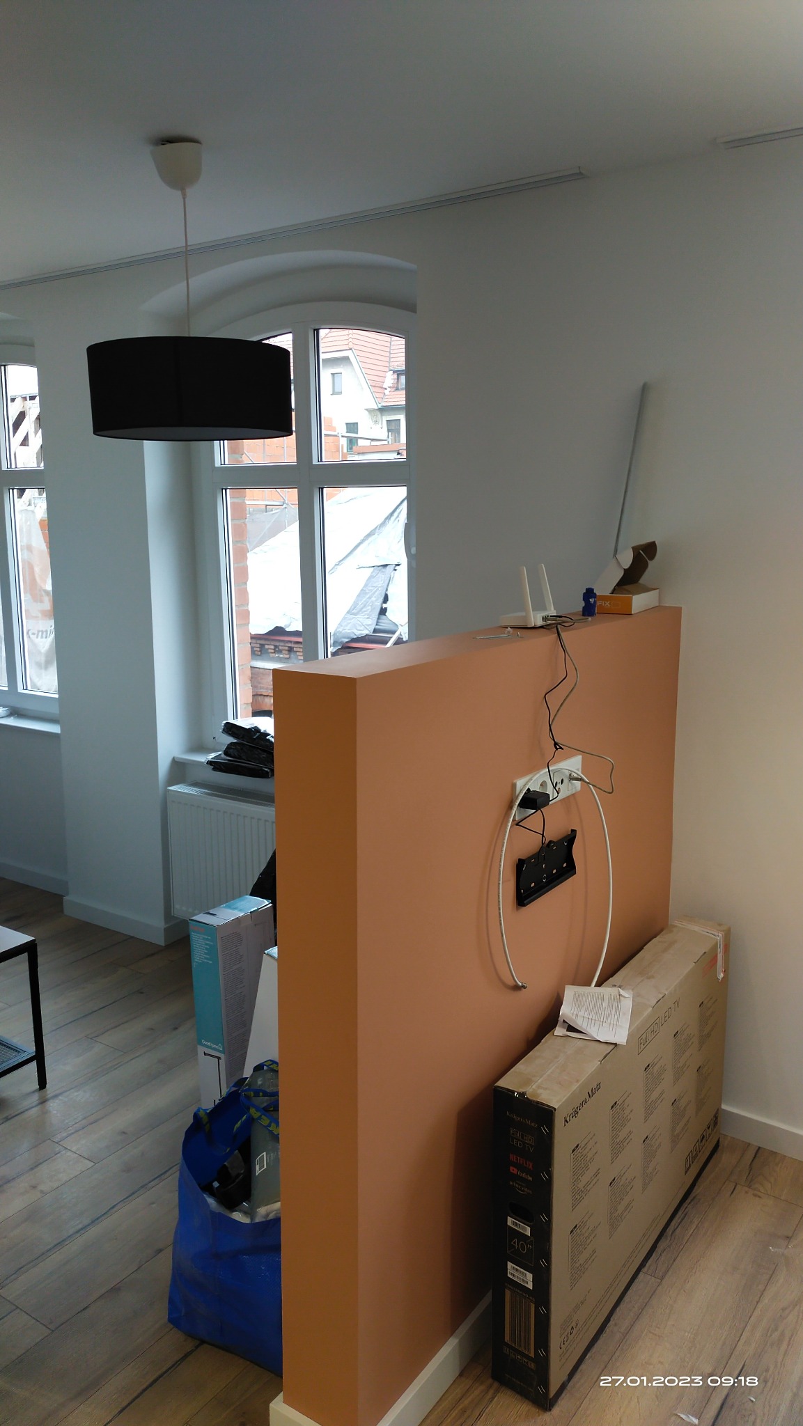 Fragment pokoju z pomarańczową ścianką działową, gniazdkiem, uchwytem na telewizor, routerem, pudełkiem po telewizorze Krüger&Matz 40 cali, niebieską torbą i lampą wiszącą.