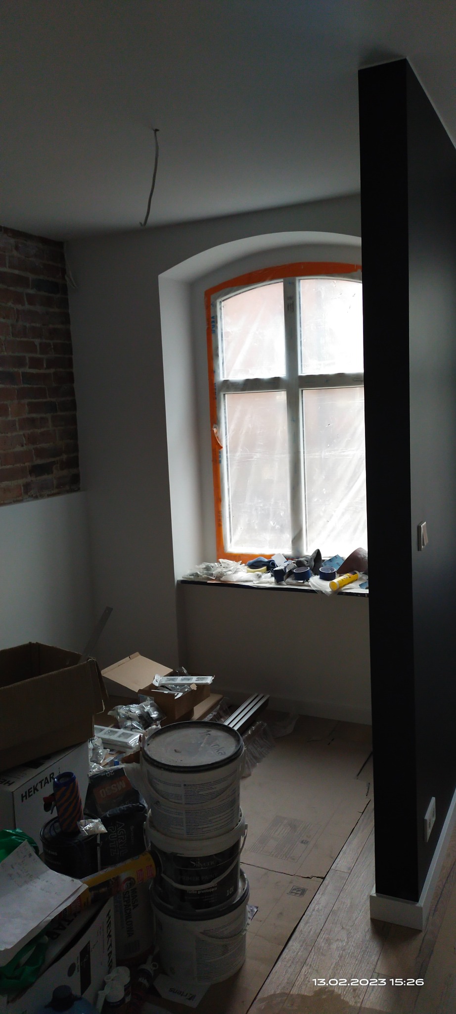 Pokój w trakcie prac wykończeniowych, widoczna ściana z cegły, okno zabezpieczone folią, puste wiadra po farbie i narzędzia na podłodze zabezpieczonej kartonem.