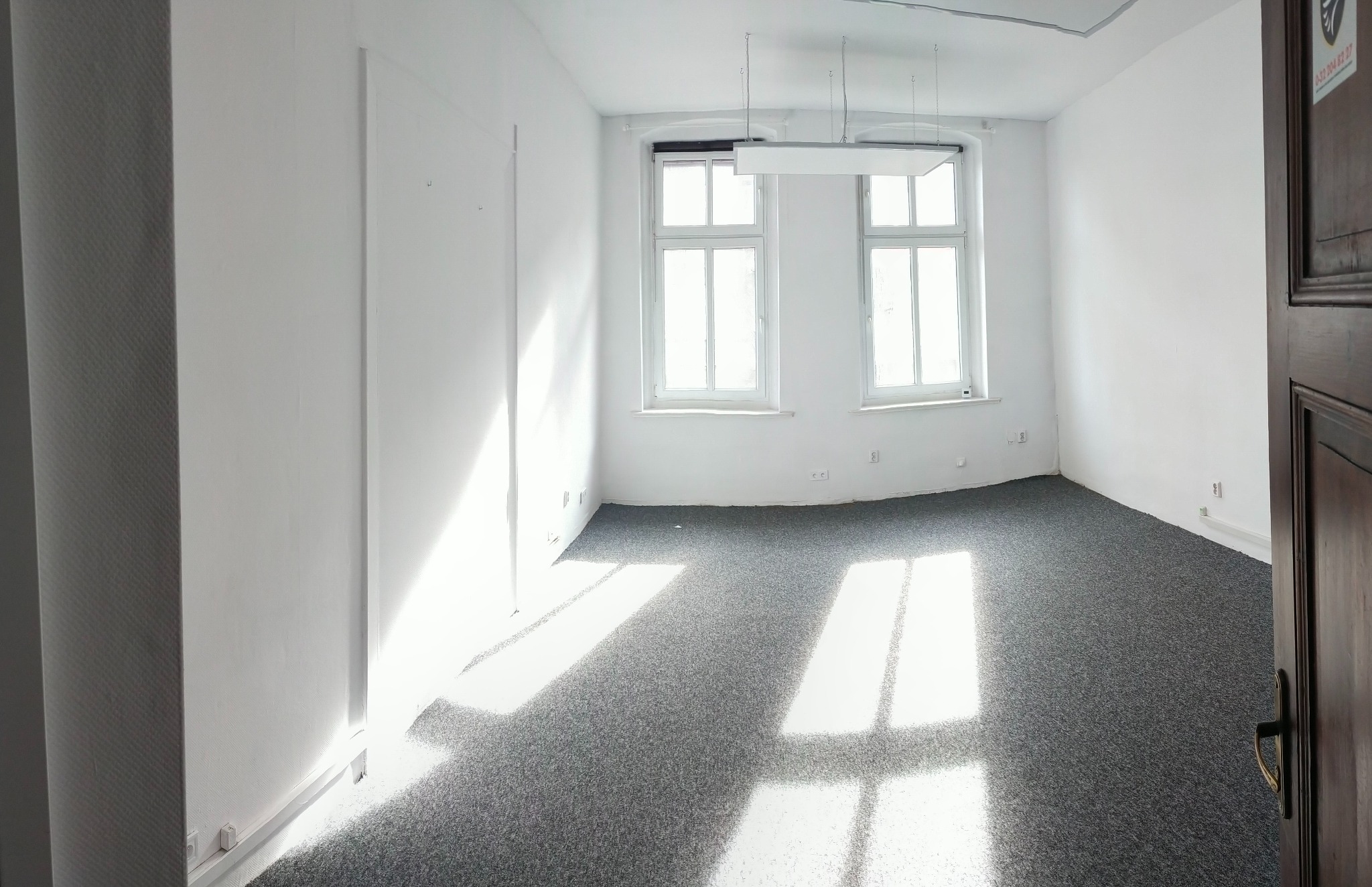 Puste, jasne pomieszczenie z szarym dywanem, dwoma oknami z białymi ramami i widoczną częścią ciemnych drzwi po prawej stronie, z logo firmy w prawym górnym rogu.