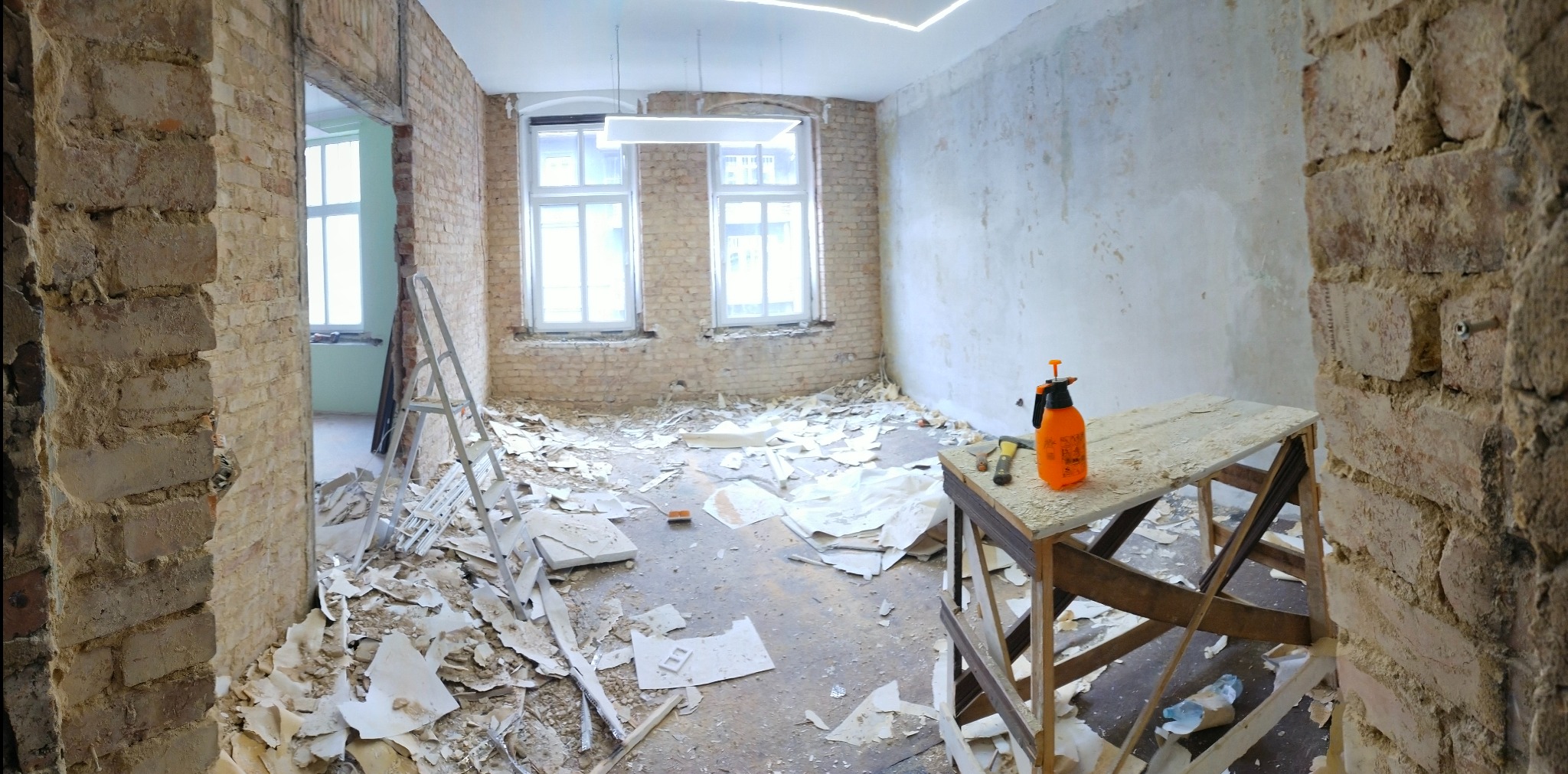 Pokój w trakcie demontażu starych powłok, widoczne odsłonięte cegły, resztki tapet na podłodze, drabina, stolik z narzędziami i spryskiwaczem, surowy stan przed dalszymi pracami wykończeniowymi.