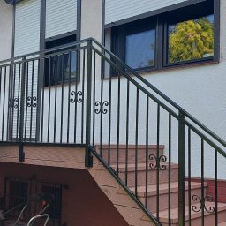 Sąsiad z Wawra - Zewnętrzne schody z kompozytu drewnopodobnego z czarną, metalową balustradą ozdobioną motywem roślinnym, prowadzące do wejścia z roletami antywłamaniowymi.