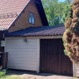 Dom w Warszawie z nowym, ciemnym dachem z blachy trapezowej, przylegający do garażu z jasną elewacją. Widoczny fragment brązowego dachu domu i fragment iglaka.