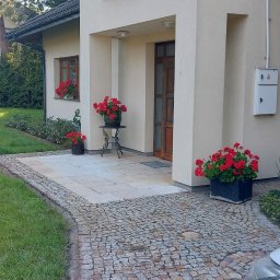 Sąsiad z Wawra - Jasna elewacja domu z drewnianymi oknami, ozdobiona czerwonymi i fioletowymi kwiatami w donicach i skrzynkach, kostka brukowa prowadząca do wejścia.