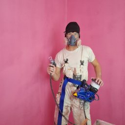 Sąsiad z Wawra - Pracownik w kombinezonie malarskim i masce ochronnej, trzymający pistolet natryskowy, na tle świeżo pomalowanej na różowo ściany w rogu pomieszczenia, zabezpieczonego folią malarską.