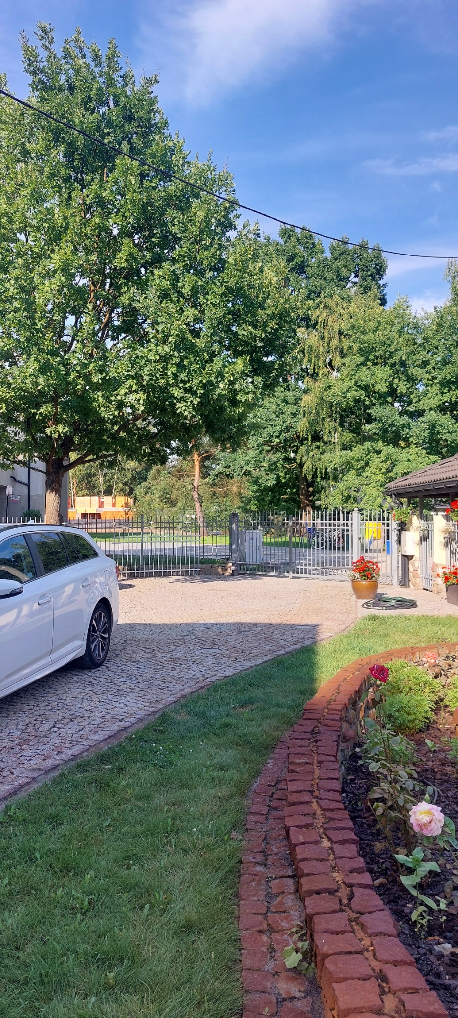 Ułożona kostka brukowa przed posesją z bramą wjazdową, widoczny fragment białego samochodu, trawnik i rabata kwiatowa