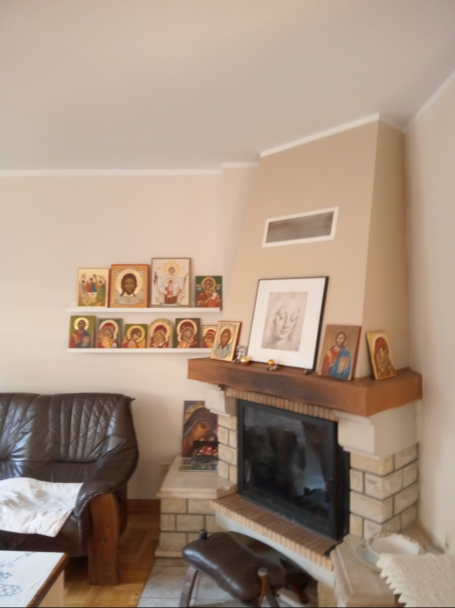 Jasne wnętrze z beżowymi ścianami, kominkiem obłożonym jasną cegłą i drewnianym blatem, nad którym wiszą obrazy sakralne i portret w ramie, obok stoi ciemnobrązowy skórzany fotel.