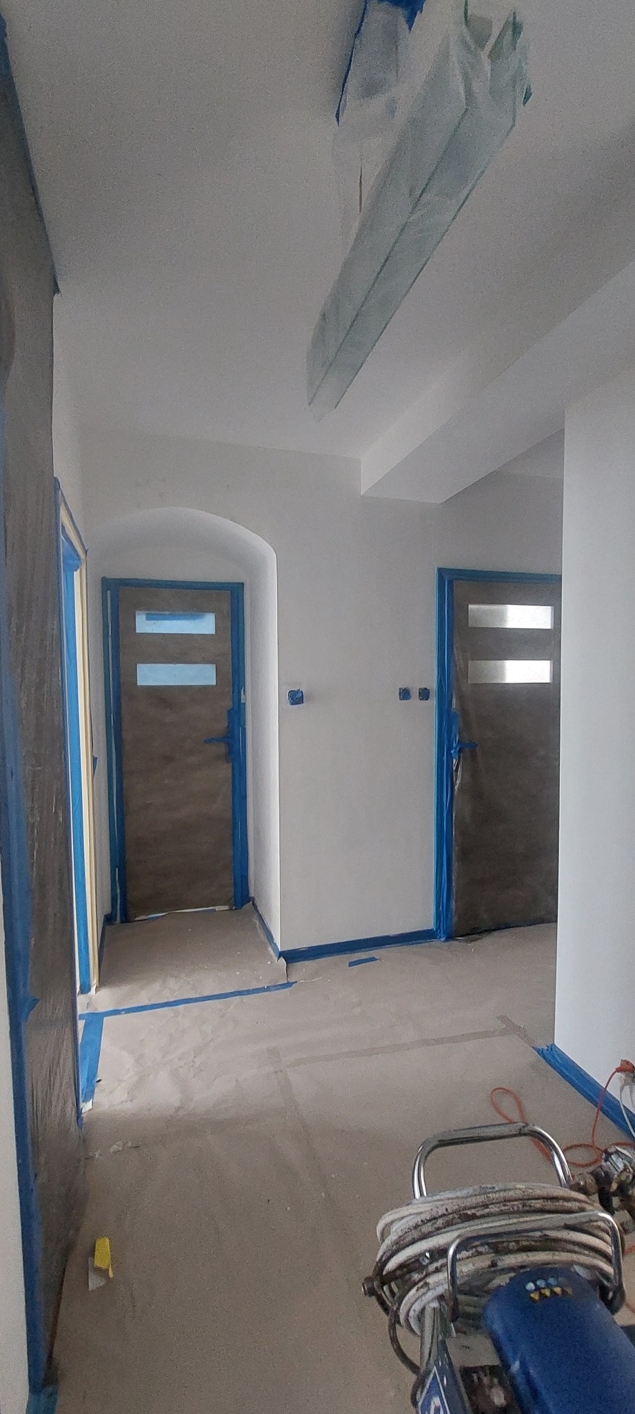 Przygotowane do malowania wnętrze z zabezpieczonymi taśmą malarską drzwiami i listwami, widoczny agregat malarski.