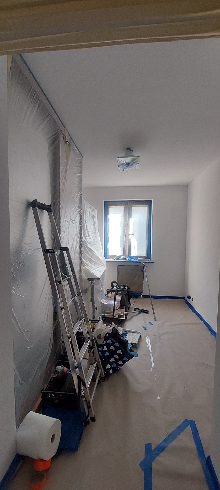 Pokój zabezpieczony folią i taśmą malarską przed odświeżeniem koloru ścian, widoczna drabina, wiaderka z farbą i akcesoria malarskie.