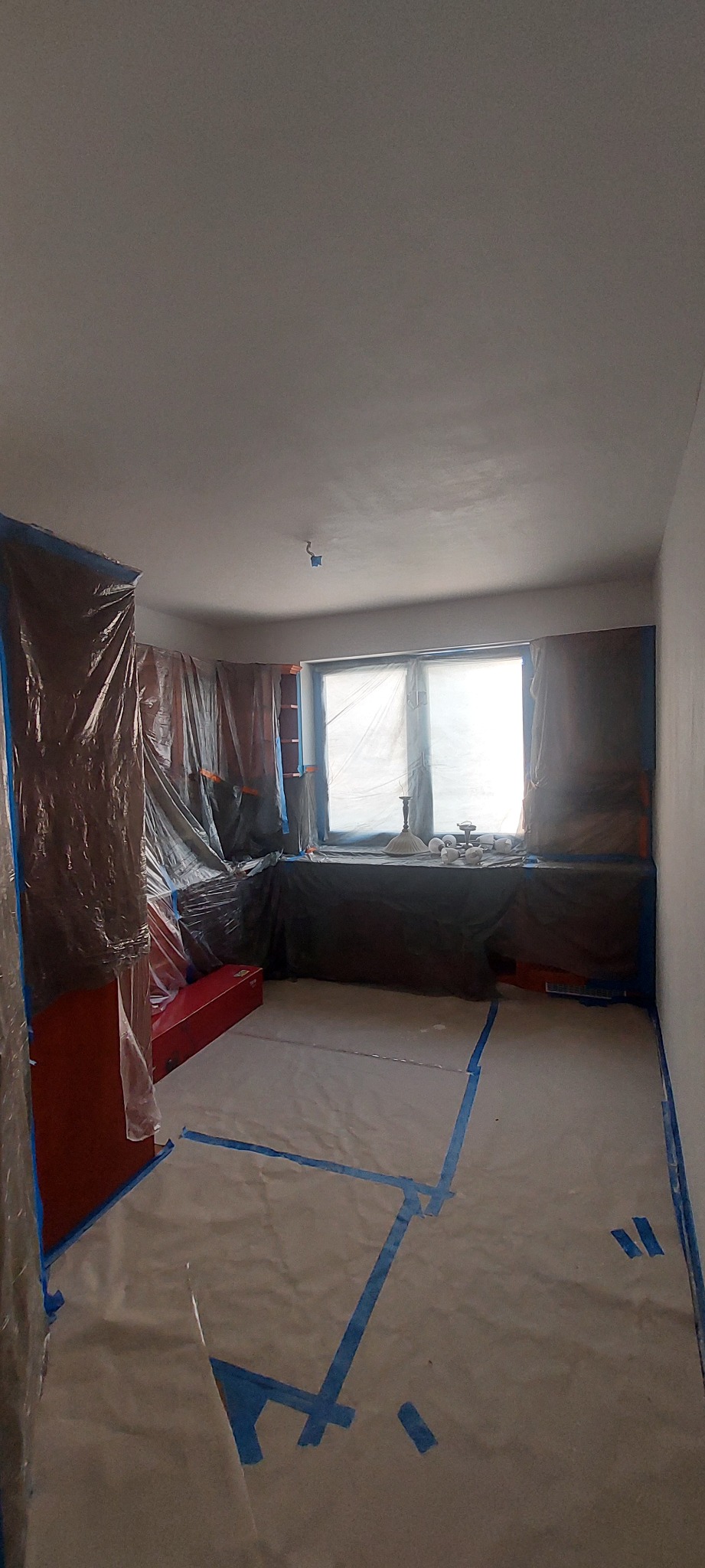 Pokój przygotowany do odświeżenia ścian: folia ochronna na meblach, oknach i podłodze, taśma malarska wyznaczająca obszar prac.