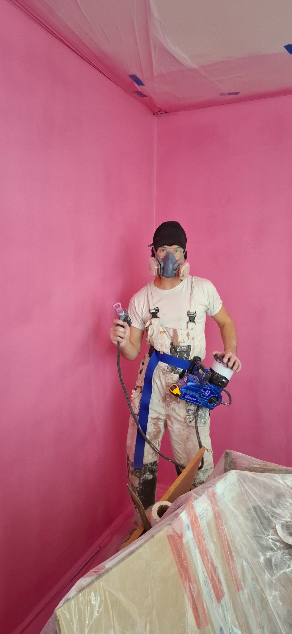 Pracownik w kombinezonie malarskim i masce ochronnej, trzymający pistolet natryskowy, na tle świeżo pomalowanej na różowo ściany w rogu pomieszczenia, zabezpieczonego folią malarską.