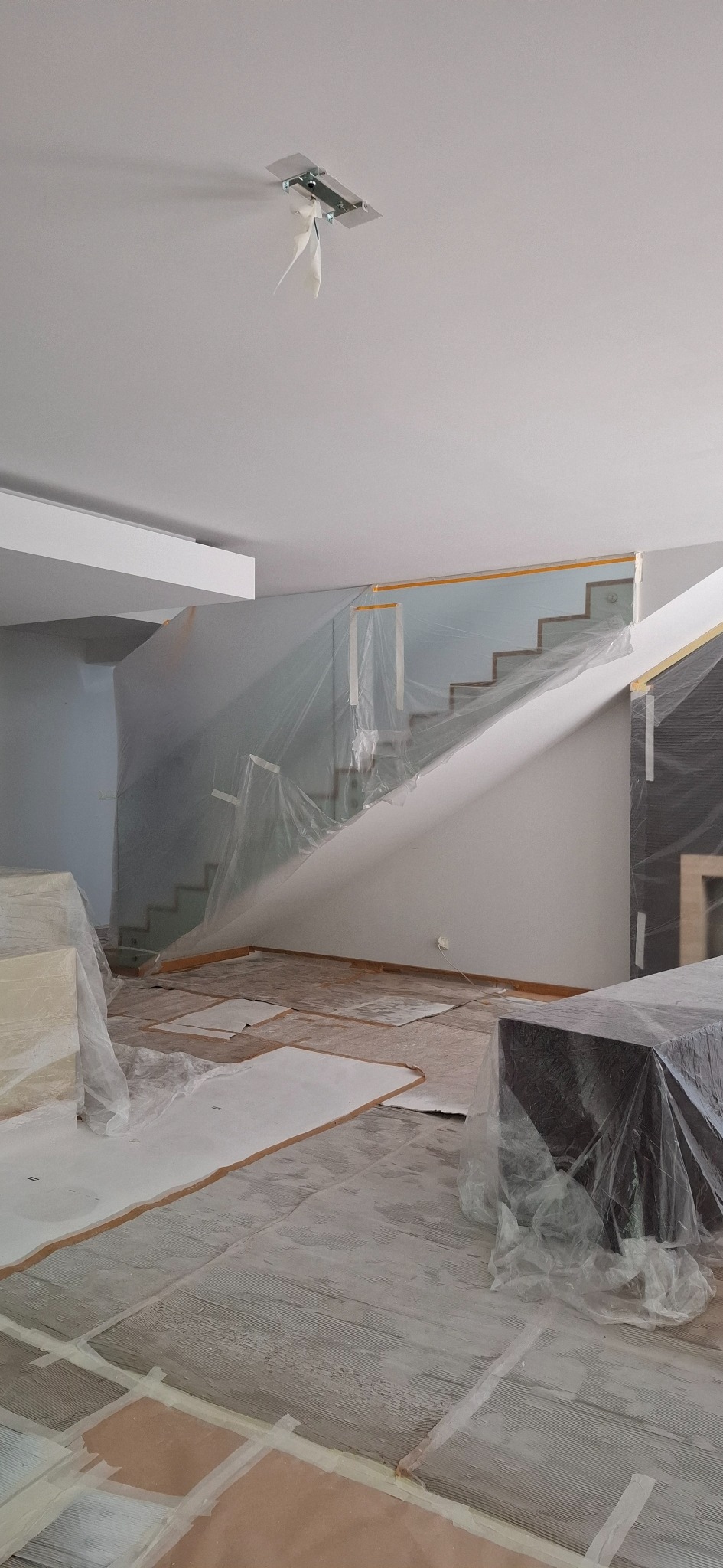 Salon w trakcie zabezpieczania folią malarską przed remontem. Widoczne schody, podłoga i meble przygotowane do prac malarskich.