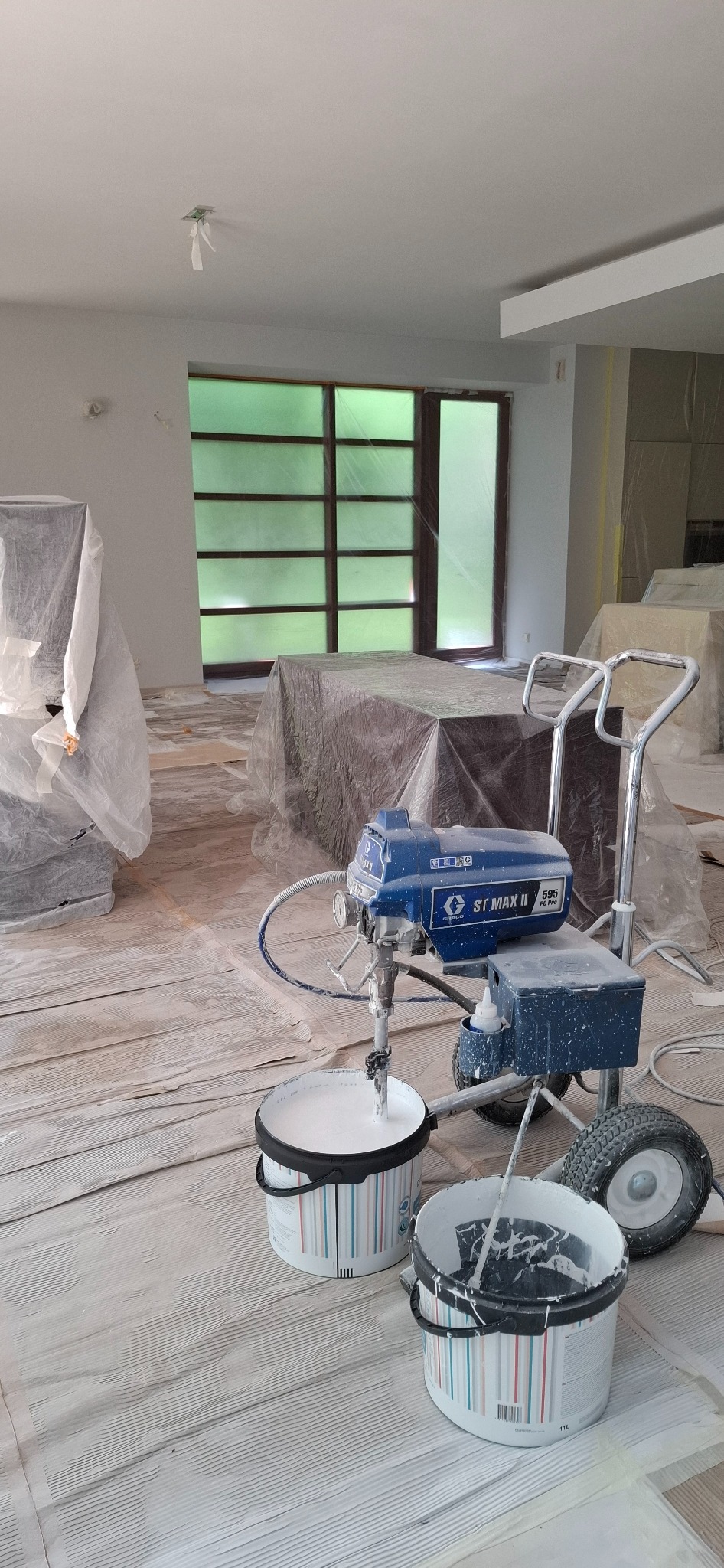 Przygotowania do malowania wnętrza: agregat malarski Graco, wiadra z farbą i folia ochronna na podłodze. Okna zabezpieczone folią malarską.
