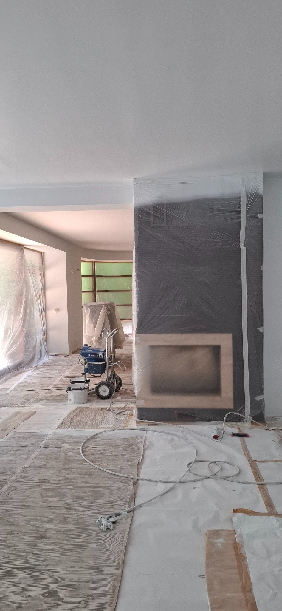 Przygotowanie do malowania wnętrza: zakryte folią okna, podłoga i kominek. Widoczny agregat malarski Wagner i wałek malarski. Ochrona przed zabrudzeniem farbą.