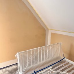 Do Ładu - Pokój w trakcie prac: ściana pomalowana na brzoskwiniowy kolor, zabezpieczone taśmą malarską połączenie ściany i sufitu, łóżko przykryte folią ochronną.