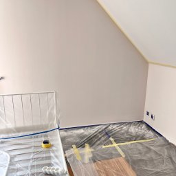 Do Ładu - Pokój w trakcie odświeżania: ściany pomalowane na jasny odcień, zabezpieczone folią podłoga i meble, widoczne taśmy malarskie na łączeniu ściany z sufitem oraz gniazdka elektryczne.