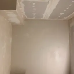 Pokój w trakcie prac wykończeniowych: ściany częściowo pokryte gładzią, widoczne ślady po szpachlowaniu na suficie i ścianach, wiadro z narzędziami na podłodze.