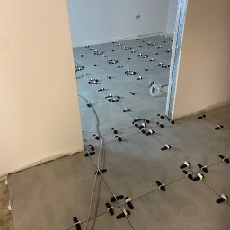 Do Ładu - Układanie szarych płytek podłogowych z czarnymi krzyżykami dystansowymi w trakcie remontu pomieszczenia, widoczne kable elektryczne na podłodze.