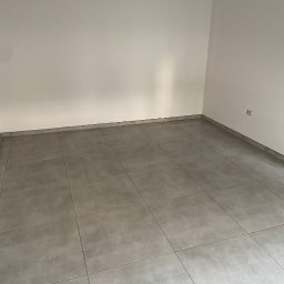 Do Ładu - Wykończone wnętrze z szarymi płytkami podłogowymi i świeżo pomalowanymi ścianami w odcieniach bieli, widoczny kontakt elektryczny na ścianie.
