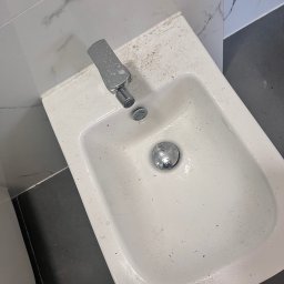 Do Ładu - Brudny bidet z chromowaną baterią i odpływem, widoczne zabrudzenia na ceramice i armaturze, tło stanowi marmurowa okładzina ścienna i ciemna podłoga.
