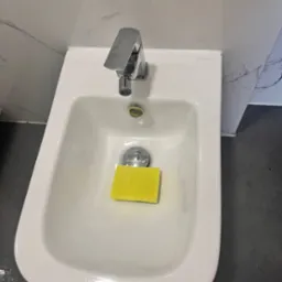 Bidet marki Omnires z chromowaną baterią i ż&oacute;łtą gąbką w środku, na tle marmurowej ściany i ciemnej podłogi.