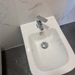 Do Ładu - Bidet z nowoczesną, chromowaną baterią, zamontowany przy ścianie wyłożonej marmurowymi płytkami. Widoczne szare fugi i ciemna podłoga z drobnymi plamami wody.