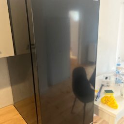 Do Ładu - Błyszcząca, czarna lodówka marki Sharp stoi w kuchni; na blacie obok widoczne akcesoria do sprzątania, w tym żółta ścierka i gąbka.