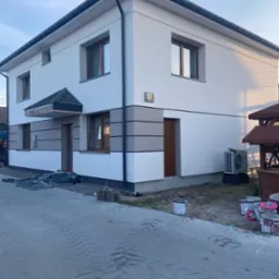 Dwukondygnacyjny dom z nową elewacją w odcieniach bieli i szarości, widoczne ślady prac malarskich, puste puszki po farbie i folia zabezpieczająca.