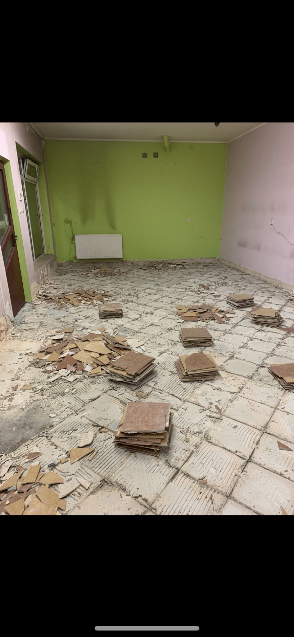 Pokój w trakcie remontu, podłoga po skuciu płytek, widoczne pozostałości kleju i fragmenty płytek, ułożone w stosy nowe płytki, ściany częściowo pomalowane na zielono i różowo.