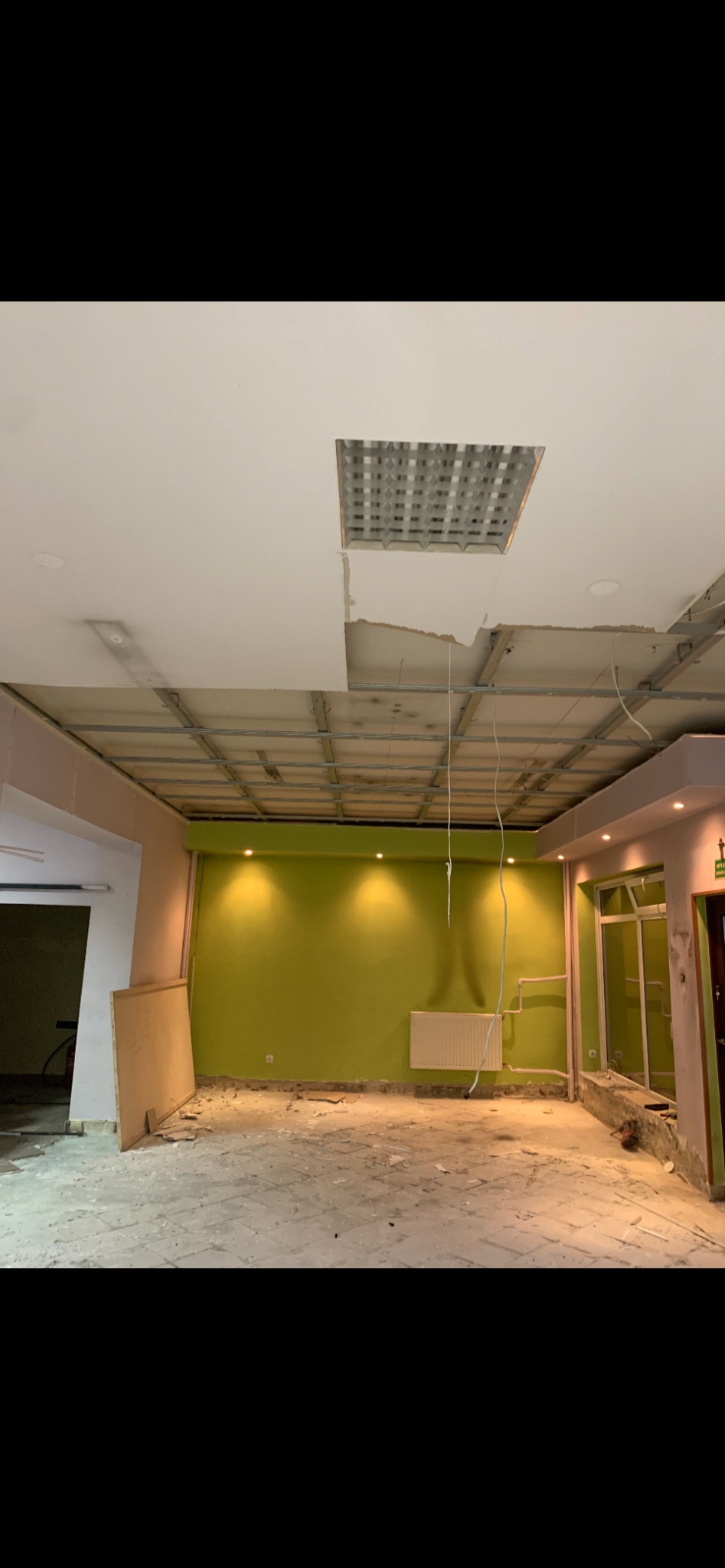 Pokój biurowy w trakcie remontu: widoczne metalowe profile sufitowe, częściowo zdemontowany sufit podwieszany, świeżo pomalowana zielona ściana i grzejnik.