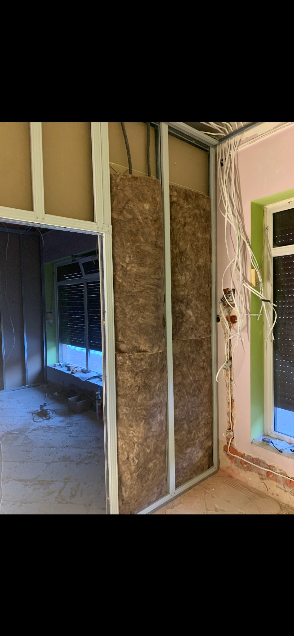 Montaż izolacji z wełny mineralnej w metalowym stelażu ściany działowej, widoczne przewody elektryczne i fragment okna z roletą.