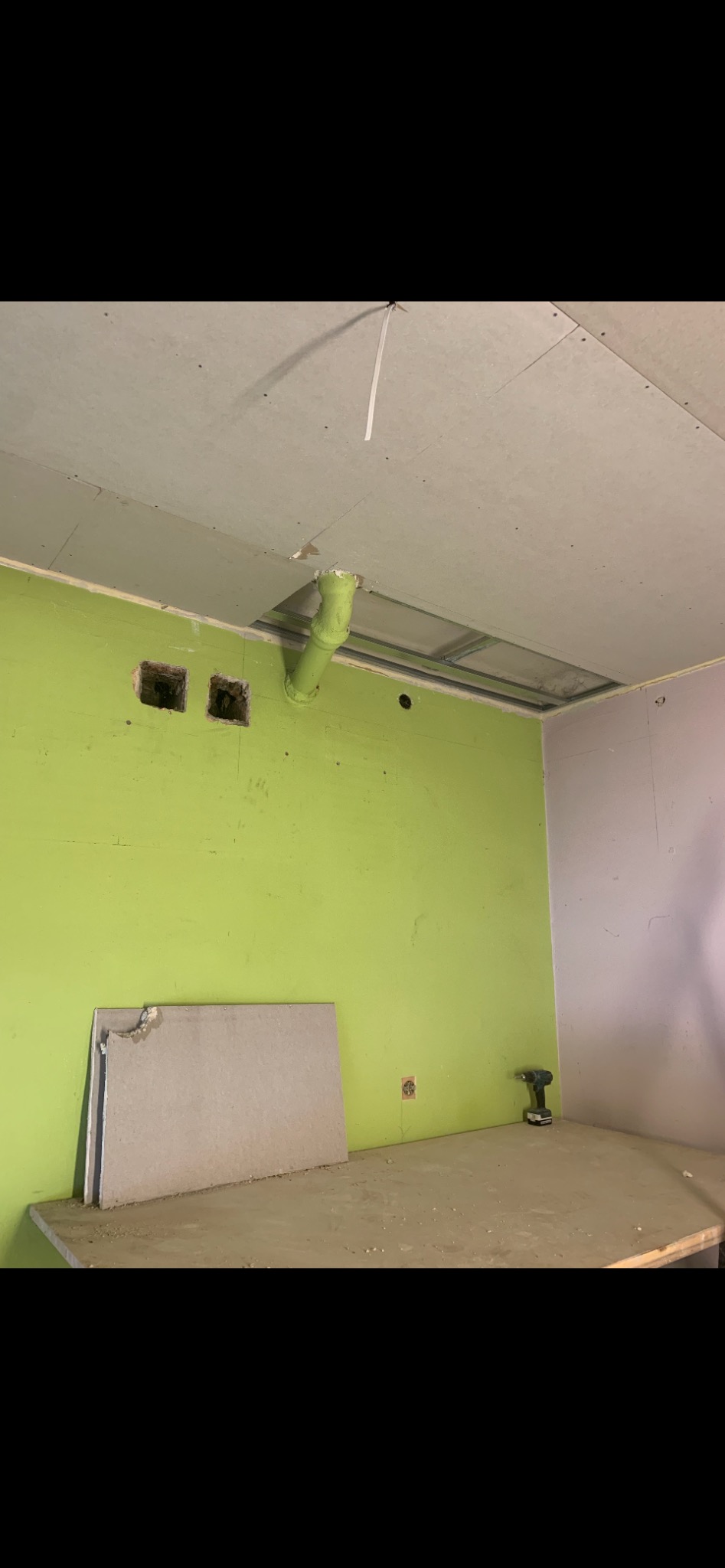 Wnętrze w trakcie prac remontowych: zielona ściana z dwoma otworami, płyty karton-gips oparte o ścianę, sufit z zamontowaną konstrukcją pod płyty i wystającą zieloną rurą, wiertarka akumulatorowa...